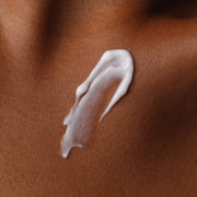 Close-up texture shot of Natural Moisturizing Factors + Inulin Body Lotion. The white, creamy product is swatched on smooth, deeply toned skin to show its rich, moisturizing consistency.